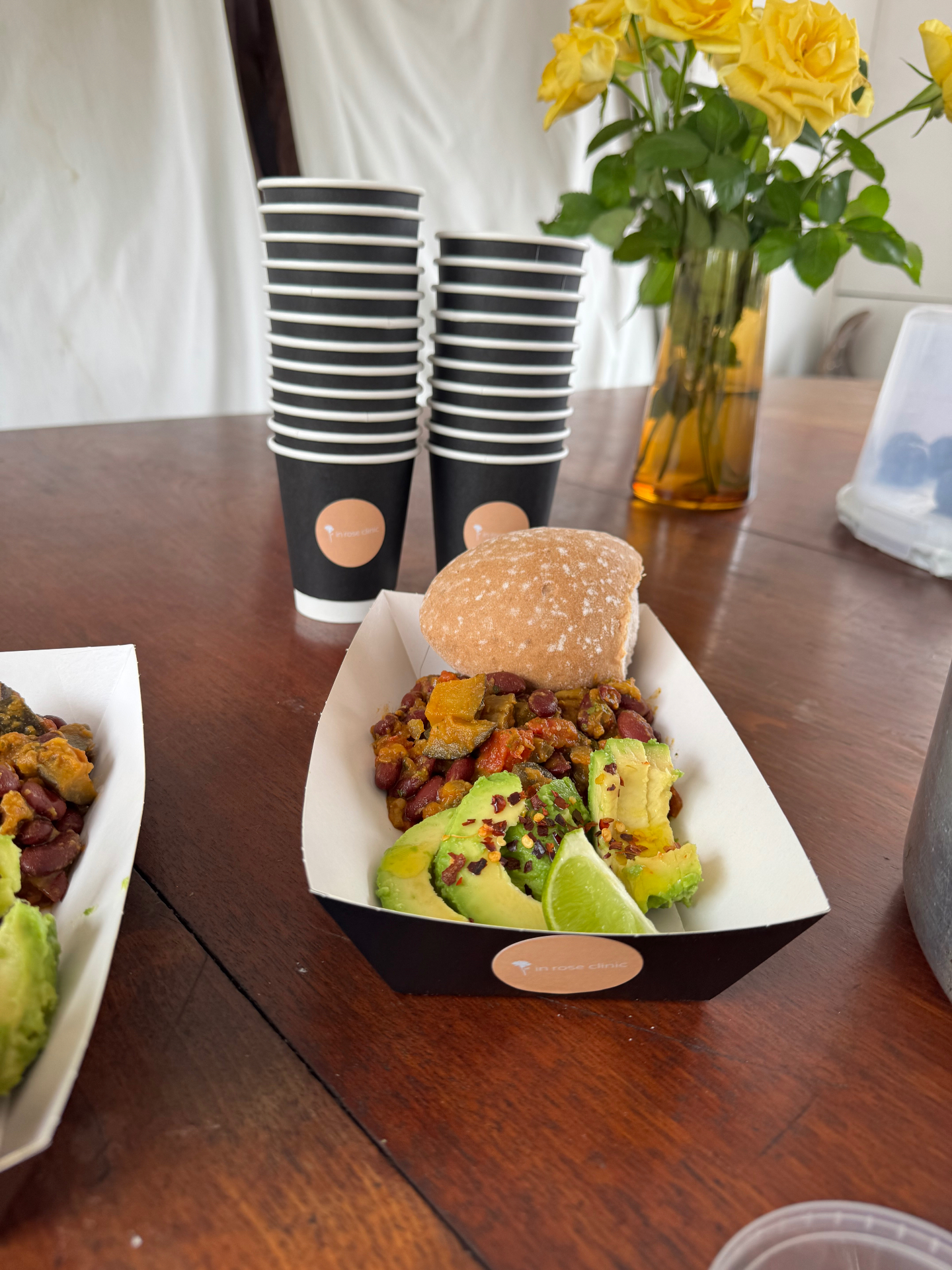 Nourishing brunch tray with bean stew, avocado, lime and a fresh roll served in branded in rose clinic packaging, beside yellow roses
